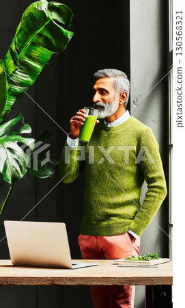 A man in a green sweater enjoys matcha while standing in a bright coworking space. Lush plants surround the area, creating a tranquil environment for productivity 130568321