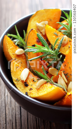 Bright orange slices of pumpkin are elegantly arranged on a dark plate with fresh garlic and rosemary alongside. This rustic display captures the essence of autumn cuisine 130568407