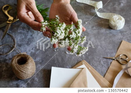 Florist at work: a pretty woman prepares a summer bouquet of white gypsophila on a gray desk. Kraft paper, scissors, greeting envelope on the table. View from above. The composition is flat. Florist at work: a pretty woman prepares a summer bouquet of white gypsophila on a gray desk. Kraft paper, scissors, greeting envelope on the table. View from above. The composition is flat. 130570442