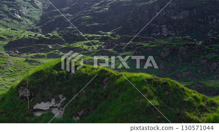 Green grass grows on rolling hills in the Gap of Dunloe, County Kerry, Ireland. Rocks are found on hillsides. The sun shines on the landscape. Green grass grows on rolling hills in the Gap of Dunloe, County Kerry, Ireland. Rocks are found on hillsides. The sun shines on the landscape. 130571044