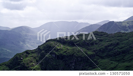 Green mountains roll into the distance in County Kerry, Ireland. The Gap of Dunloe vista is shrouded in mist and low-hanging clouds, creating a tranquil and peaceful atmosphere. Green mountains roll into the distance in County Kerry, Ireland. The Gap of Dunloe vista is shrouded in mist and low-hanging clouds, creating a tranquil and peaceful atmosphere. 130571050