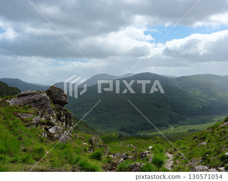 A vista across the Gap of Dunloe in Ireland shows lush green hills and valleys under a cloudy sky. Rock formations are visible on the hillside in the foreground. 130571054