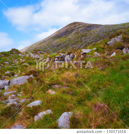 A picturesque view of a hiking path with grass and rocks, leading up towards majestic mountains under a sunny blue sky with scattered clouds in Gap of Dunloe, Ireland. 130571058