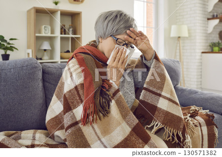 Sick upset senior woman with runny nose wrapped in a blanket sitting at home. Sick upset senior woman with runny nose wrapped in a blanket sitting at home. 130571382