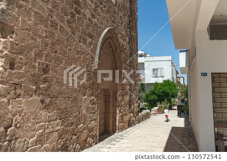 Tourists walking narrow stone-walled street with pointed arch, revealing medieval architectural heritage of Famagusta's historic urban landscape. Cyprus 130572541