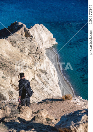Photographer hiking white limestone cliffs of pissouri, carrying backpack, overlooking azure mediterranean waters under bright sunlight 130572545