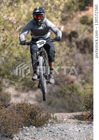 Mountain biker wearing protective equipment airborne during high-speed trail jump surrounded by lush wilderness landscape 130572560
