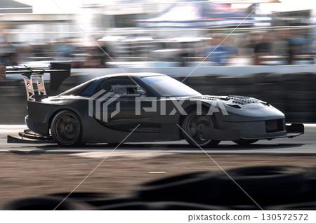Racing sports car gleaming with dark gray paint, blurring against sunlit racetrack while accelerating with powerful momentum 130572572
