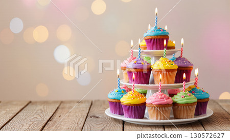 Colorful cupcakes with birthday candles and sprinkles arranged on tiered stand over wooden table with soft pastel bokeh background. 130572627
