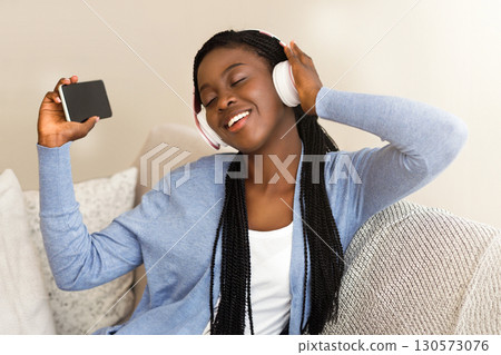 Favourite Song. Cheerful African American Girl In Headphones Listening Music On Smartphone And Singing On Sofa At Home 130573076
