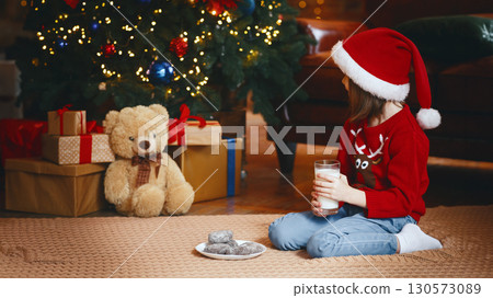 Preschool girl in red Santa hat holding glass of milk, looking at teddy bear, playing with toys on Christmas Eve by xmas tree 130573089