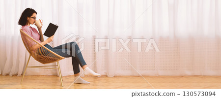 Free Time. Young woman in glasses reading book sitting in chair by the window on spring morning, drinking tea, copyspace Free Time. Young woman in glasses reading book sitting in chair by the window on spring morning, drinking tea, copyspace 130573094