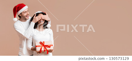 Personal Santa. Loving Boyfriend Making Christmas Surprise to His Black Girlfriend, Covering Her Eyes And Presenting Gift, Yellow Studio Background Personal Santa. Loving Boyfriend Making Christmas Surprise to His Black Girlfriend, Covering Her Eyes And Presenting Gift, Yellow Studio Background 130573129