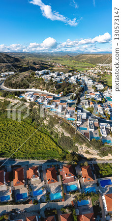 Aerial view of Pissouri village in Cyprus, featuring terraced houses with private pools amid lush green fields and hills under clouds 130573147