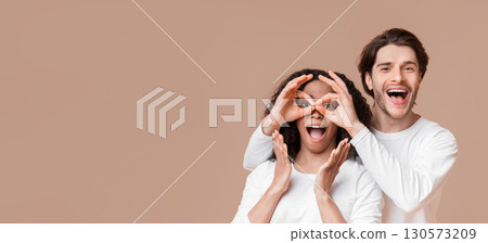 Cheerful multiracial couple having fun, fooling together. Man making funny glasses with fingers to his girlfriend over yellow background 130573209