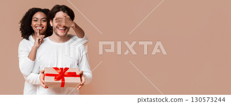 Surprise gift. Afro girlfriend covering her boyfriend's eyes and showing silence sign while giving birthday or Christmas present, yellow background 130573244