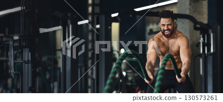 A muscular man energetically utilizes battle ropes in a gym, demonstrating strength and focus as he engages in a vigorous workout routine, copy space A muscular man energetically utilizes battle ropes in a gym, demonstrating strength and focus as he engages in a vigorous workout routine, copy space 130573261