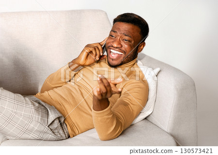 Joyful African American Guy Talking On Cellphone And Laughing Holding Laptop Computer Lying On Sofa Indoor. Selective Focus Joyful African American Guy Talking On Cellphone And Laughing Holding Laptop Computer Lying On Sofa Indoor. Selective Focus 130573421