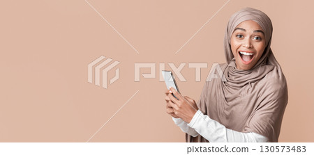 Unexpected message. Portrait of excited afro muslim girl in hijab holding smartphone and looking at camera, yellow background with free space 130573483