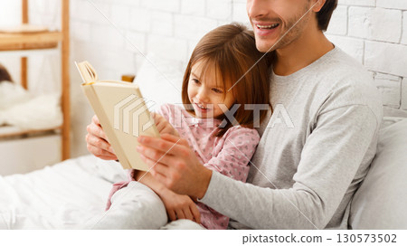 Close up of preschool cute girl reading book with father, sitting on bed together Close up of preschool cute girl reading book with father, sitting on bed together 130573502