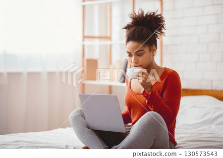 Weekend Morning. Relaxed Black Woman Using Laptop Having Coffee Sitting On Bed In Bedroom. Copy Space, Selective Focus Weekend Morning. Relaxed Black Woman Using Laptop Having Coffee Sitting On Bed In Bedroom. Copy Space, Selective Focus 130573543