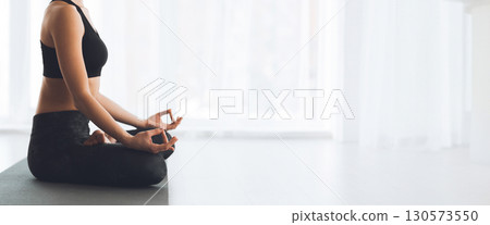 A woman sits in a cross-legged position on a mat, practicing meditation with a calm expression in a bright, peaceful environment, copy space 130573550
