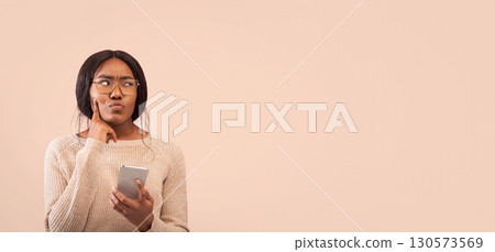Pensive Afro Girl Holding Cellphone Thinking About Something Touching Chin Standing On Pink Background. Studio Shot, Empty Space 130573569