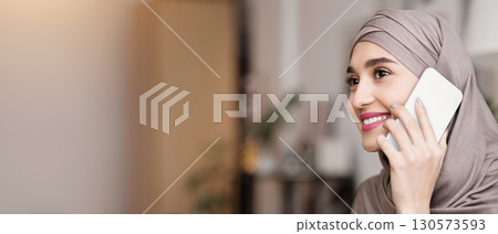 In Touch. Closeup portrait of modern muslim woman in hijab talking on cellphone indoors, smiling and looking away, selective focus In Touch. Closeup portrait of modern muslim woman in hijab talking on cellphone indoors, smiling and looking away, selective focus 130573593