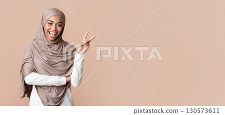 Cheerful black muslim girl in hijab showing peace gesture or v-sign and looking at camera, posing over yellow studio background, free space 130573611