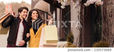A couple smiles at each other while holding shopping bags and a credit card, walking along a bustling city street in autumn, copy space 130573669