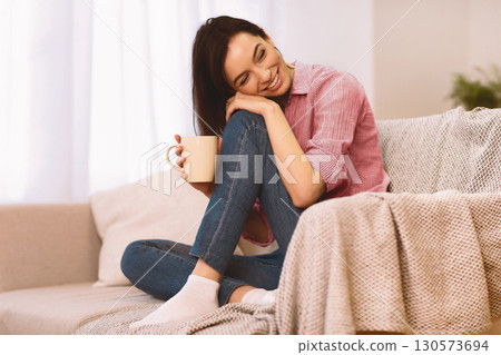 Side view portrait of happy young woman relaxing at home sitting on a sofa in the living room with a cup of coffee Side view portrait of happy young woman relaxing at home sitting on a sofa in the living room with a cup of coffee 130573694