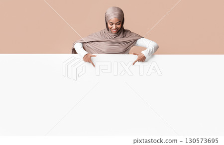 Free Space For Your Ad. Black muslim woman in headscarf looking down and pointing at blank white advertisement board over yellow studio background Free Space For Your Ad. Black muslim woman in headscarf looking down and pointing at blank white advertisement board over yellow studio background 130573695
