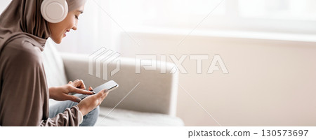 Joyful arabic girl in hijab listening music, wearing wireless headphones and using mobile phone at home, selective focus, crop 130573697