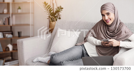 Cheerful muslim girl in hijab spending time with smartphone at home and looking at camera, enjoying lazy weekend. 130573700