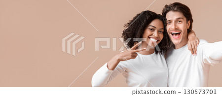 Cheerful millennials taking selfie. Joyful interracial couple capturing self-portrait, posing together over yellow background, girl gesturing peace 130573701