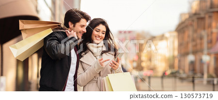 Cheerful couple shopaholics checking info about season sales on mobile phone, walking by street together, holding purchases, copy space 130573719