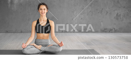 A woman sits in prayer pose on a mat, reflecting a calm demeanor in a serene studio illuminated by soft morning light, copy space 130573758