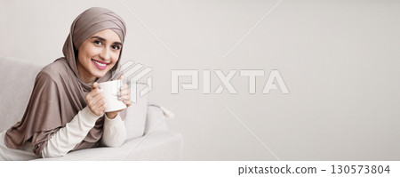 Portrait of happy muslim girl in hijab relaxing at home with cup of coffee, sitting on sofa and looking at camera, copy space Portrait of happy muslim girl in hijab relaxing at home with cup of coffee, sitting on sofa and looking at camera, copy space 130573804