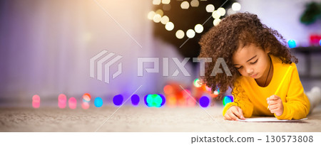 Cherished wish. Focused little african girl drawing letter to Santa Claus, lying on floor near Christmas tree, empty space 130573808
