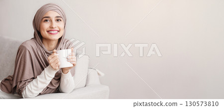 Happy arabic womanin hijab relaxing on couch with cup of tea, enjoying weekend at home and looking at camera, copy space 130573810