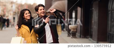 Wow sales. Guy pointing on shopwindow, shopping with wife in modern boutiques outdoors, copy space Wow sales. Guy pointing on shopwindow, shopping with wife in modern boutiques outdoors, copy space 130573912