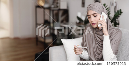 Sick arabic girl in hijab holding thermometer and calling family doctor to get consultation, sitting on couch at home 130573913