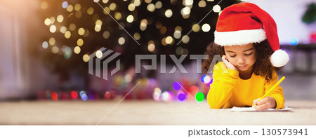 Focused little girl writing letter to Santa Claus, lying on floor near Christmas tree 130573941