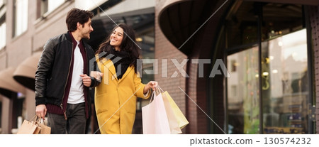 A couple walks hand in hand outside a store, carrying shopping bags and smiling at each other, copy space A couple walks hand in hand outside a store, carrying shopping bags and smiling at each other, copy space 130574232