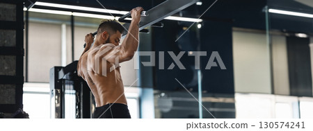 A man exercises by doing pull-ups at a well-equipped gym, focusing on building upper body strength, copy space 130574241