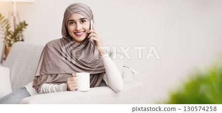 Happy Arabic Girl In Headscarf Calling To Friend And Drinking Coffee At Home, Relaxing On Couch In Living Room And Looking At Camera, Free Space 130574258