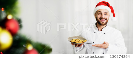 Christmas Cookies Recipes. Baker Man In Santa Hat Holding Plate With Baked Biscuits Standing Over White Background Indoor. Selective Focus 130574264