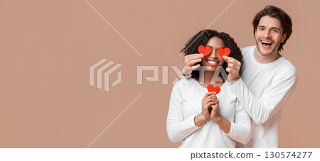 Falling In Love. Cheerful Man Holding Red Paper Hearts Over Eyes Of His Afro Girlfriend And Smiling At Camera, Yellow Background With Empty Space Falling In Love. Cheerful Man Holding Red Paper Hearts Over Eyes Of His Afro Girlfriend And Smiling At Camera, Yellow Background With Empty Space 130574277