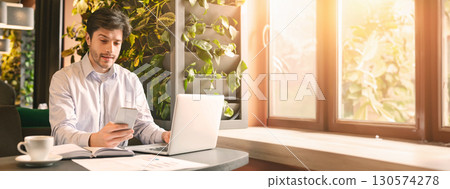 A man sits at a desk using a smartphone while working on a laptop, surrounded by plants and sunlight streaming through the window, copy space A man sits at a desk using a smartphone while working on a laptop, surrounded by plants and sunlight streaming through the window, copy space 130574278