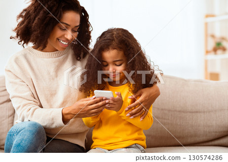 Happy Afro Mother And Daughter Using Mobile Phone Playing Games Together Sitting On Sofa Indoor. Copy Space Happy Afro Mother And Daughter Using Mobile Phone Playing Games Together Sitting On Sofa Indoor. Copy Space 130574286